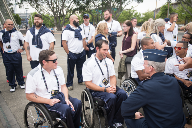 Eine Gruppe von Menschen im Rollstuhl, die vor einer Kulisse aus Bäumen, Pfählen, Gebäuden und einem klaren blauen Himmel diskutieren, wobei einige Mützen und Schutzbrillen tragen, was darauf hindeutet, dass sie bei den Paralympischen Spielen sind.