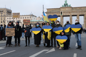 Eine Gruppe von Menschen mit Masken und Fahnen und Schildern steht vor dem Brandenburger Tor in Berlin, Deutschland, mit Gebäuden, Laternenmasten, Ampeln und einem Bogen mit Statuen im Hintergrund.