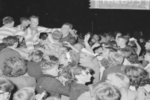 Ein Schwarz-Weiß-Foto einer Menge bei einem Konzert mit einer Gruppe von Männern, Frauen und einem Mädchen in der Mitte und Text in der rechten oberen Ecke.