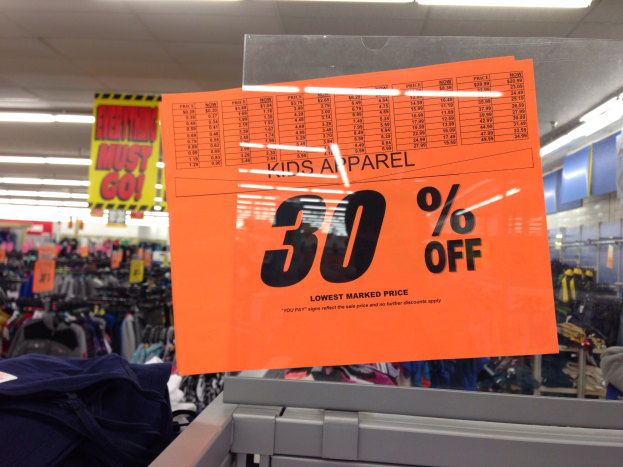 Ein Geschäft mit einem Schild "Kids Apparel 30% Off" im Vordergrund, Kleider im Hintergrund und ein Textbrett auf der rechten Seite.