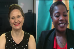 Zwei Frauen, die während eines Videoanrufs lächeln, eine in einem schwarzen und weißen Kleid, die andere in einer unbestimmten Farbe, mit einer Flagge an der Wand hinter ihnen in einer öffentlichen Einrichtung.