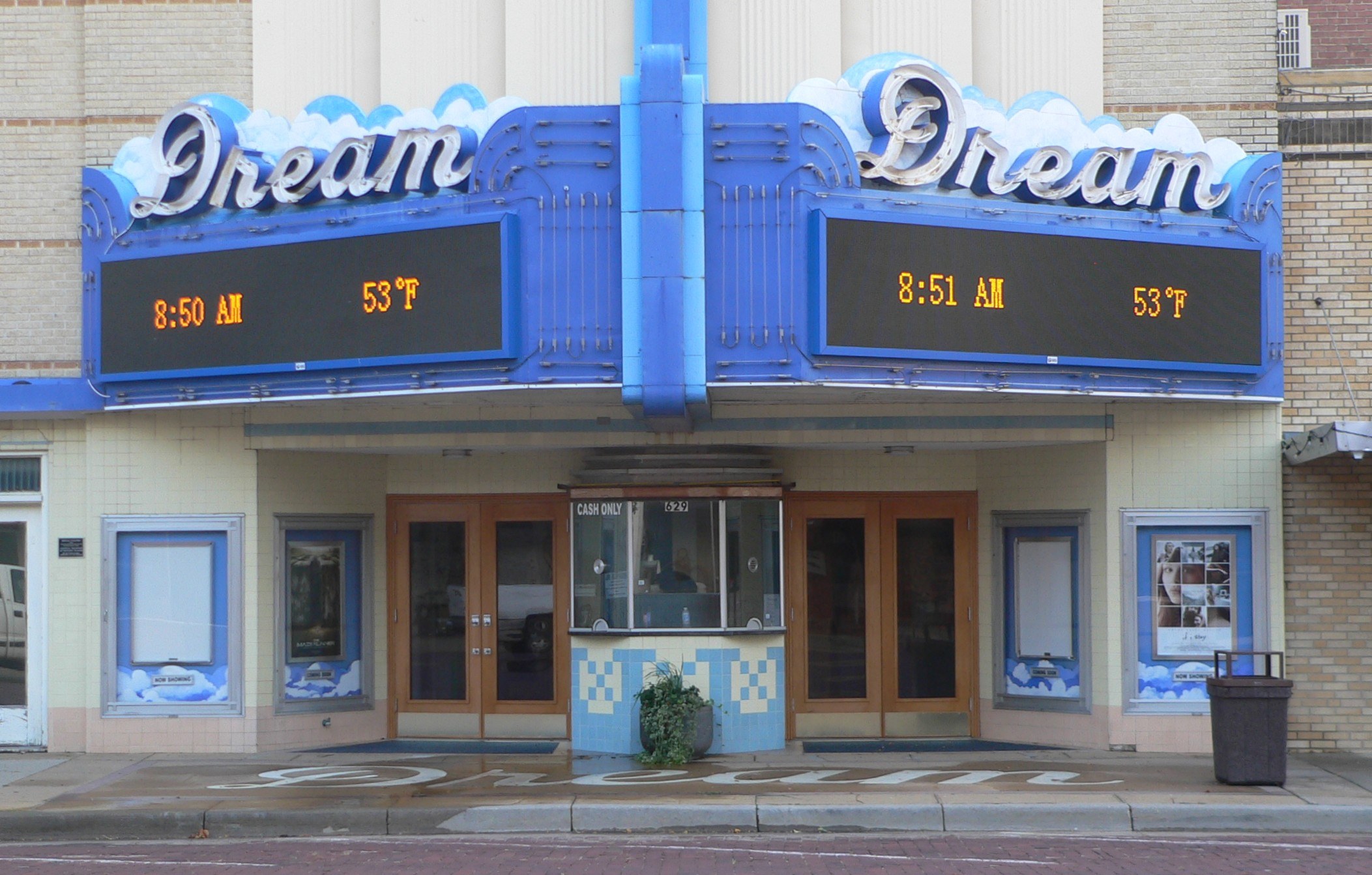 Außenansicht des Dream Theaters in St. Louis, Missouri, mit Glastüren und -fenstern, einem Namensschild, einem Müllbehälter, einem Topf mit einer Pflanze und einer Straße unten.