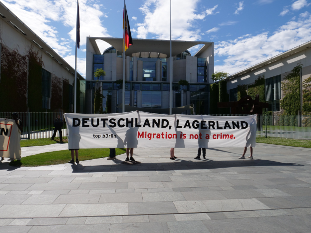Eine Gruppe von Menschen hält ein Transparent mit der Aufschrift "Deutschland, Lagerland Migration ist kein Verbrechen" vor einem Gebäude mit Fenstern, umgeben von Gras, einem Metallzaun, Pflanzen, Bäumen und Fahnen auf Stangen unter einem bewölkten Himmel.