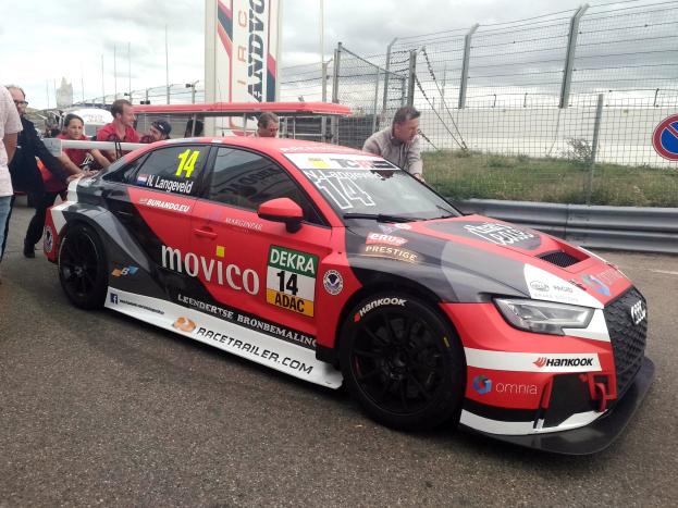 Audi RS3 DTM Rennauto auf einer Straße umgeben von Menschen, mit einem Zaun, Gras, einem Schild, einer Fahne, Pfosten und einem bewölkten Himmel im Hintergrund.