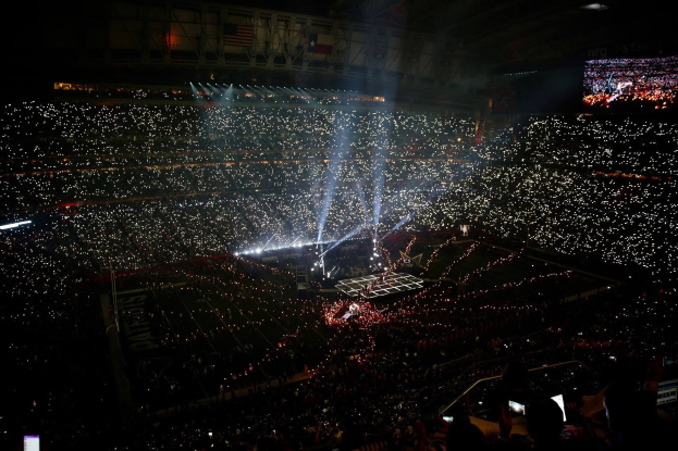 Eine große Menge in einem hell erleuchteten Stadion bei Nacht schaut auf einen Bildschirm mit den Ergebnissen der Super Bowl XLVII-Halbzeitshow.