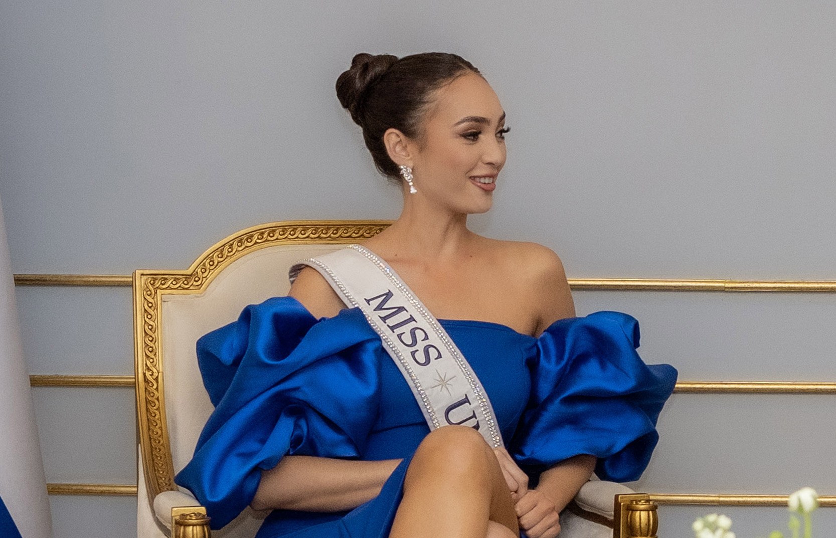 Frau in einem blauen Kleid mit einer "Miss Universe 2019"-Schärpe, die auf einem Stuhl sitzt und lächelt.