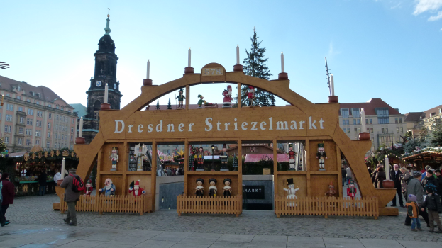 Ein belebter Weihnachtsmarkt in Dresden, Deutschland, mit Menschen, die spazieren gehen, einige mit Taschen, unter einem Bogen, mit Bäumen und Gebäuden im Hintergrund unter einem bewölkten Himmel.