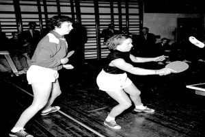 Zwei Frauen beim Tischtennis auf einem Court, jede hält einen Schläger in der Hand, mit einigen Zuschauern im Hintergrund und einem weiteren Tischtennistisch auf der rechten Seite, dargestellt in Schwarz-Weiß.