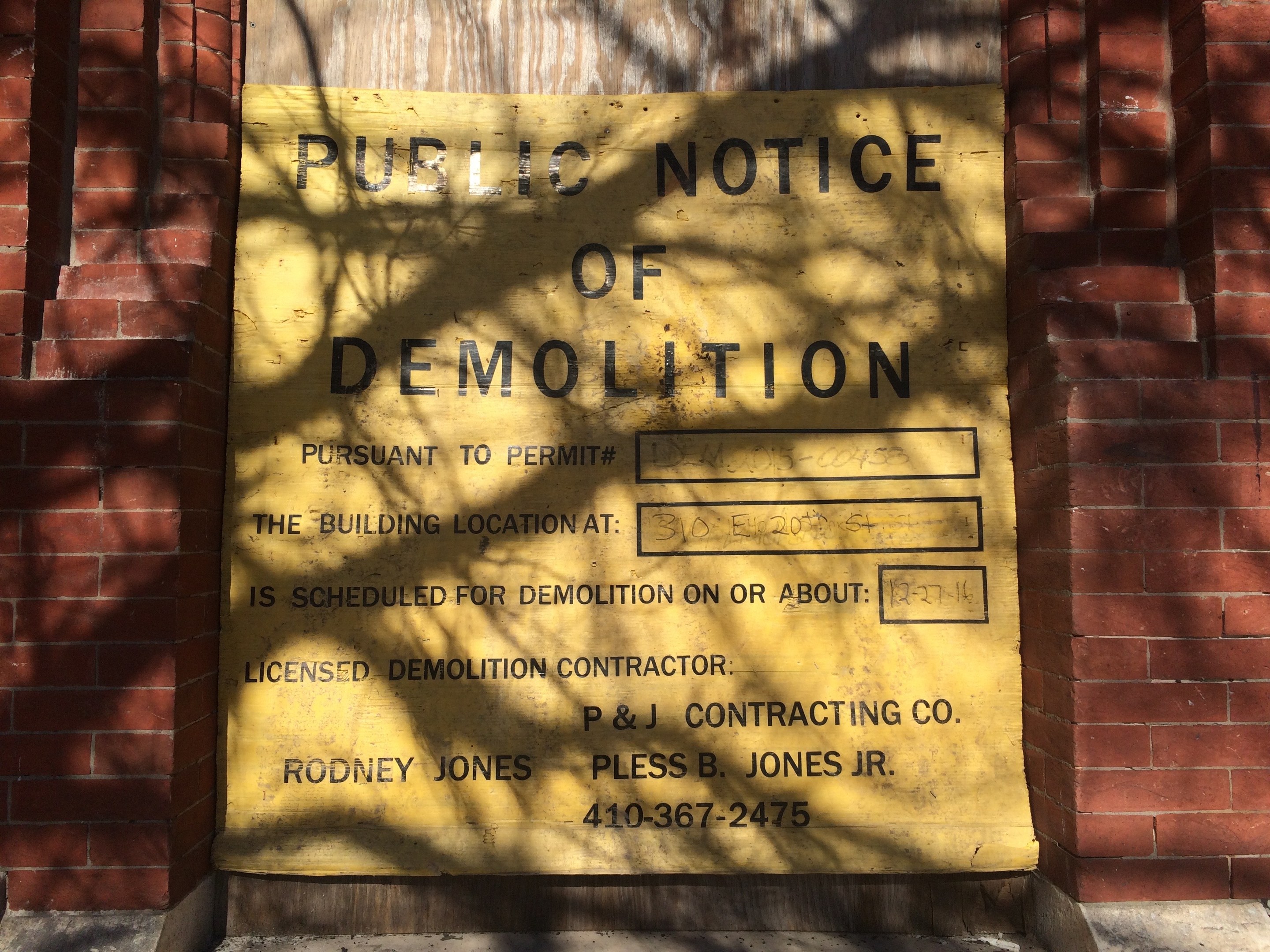 Gelbes Schild auf einem Backsteingebäude mit der Aufschrift "Öffentliche Bekanntmachung der Abriss" an der Wand angebracht.