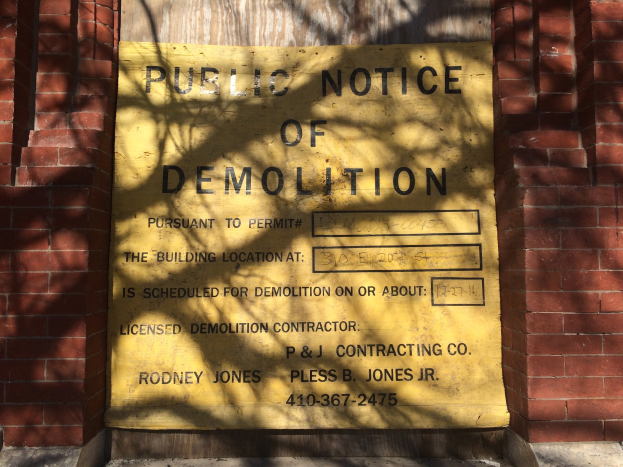 Gelbes Schild auf einem Backsteingebäude mit der Aufschrift "Öffentliche Bekanntmachung der Abriss" an der Wand angebracht.