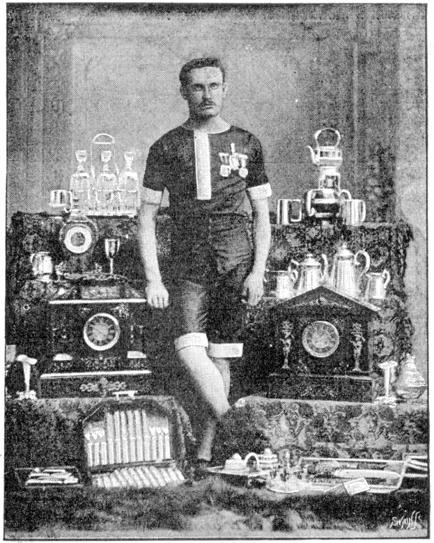 Schwarzes-Weiß-Foto eines Mannes in einem Fußballtrikot, der vor einem Tisch mit Pokale, Trophäen und anderen Gegenständen steht, mit Text unten: "1909-1910 Weltcup-Sieger."