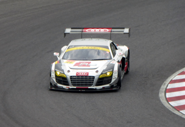 Audi R8 LMS GT3 Rennwagen in leuchtendem Rot und Weißer Lackierung, der auf einer Rennstrecke mit dem Fahrer in schwarzem Rennanzug und Helm fährt.