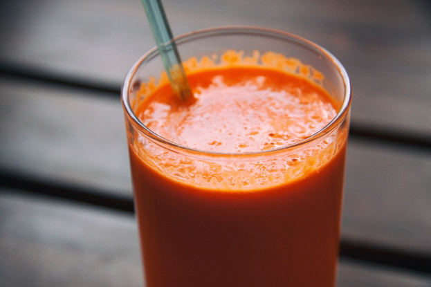 Ein Glas randvoll mit leuchtend orangefarbenem Karottensaft, ein Strohhalm darin, auf einem hell holzfarbenen Tisch mit einem sanft unscharfen Hintergrund gestellt.
