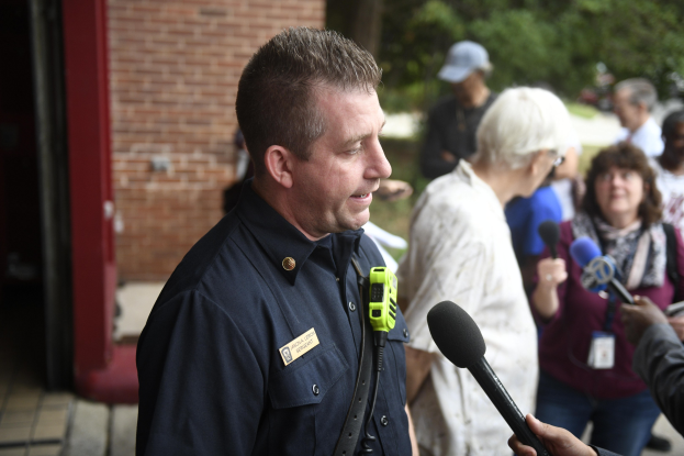 Polizeibeamter spricht mit Reportern mit Mikrofonen vor Bäumen und einem Gebäude.