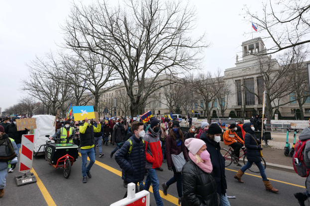 Eine große Gruppe von Menschen marschiert auf einer Straße in Washington, D.C., mit Plakaten und Schildern, einige fahren Fahrröder, unter einem klaren blauen Himmel.