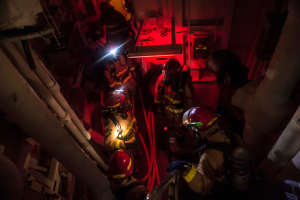 Feuerwehrleute in Helmen und Sauerstoffflaschen stehen in einem dunklen Raum mit Rohren auf der linken und rechten Seite, mit einer Maschine und anderen Gegenständen im Hintergrund, was auf einen Rettungseinsatz hinweist.
