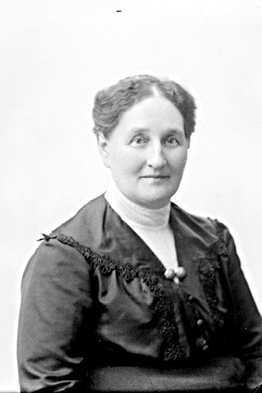 Schwarz-weißes Porträt einer selbstbewussten Frau in einem schlichten, eleganten Kleid mit langen Ärmeln und hohem Kragen, deren Haar zurückgebunden ist und die direkt in die Kamera blickt, mit einem warmen, bestimmten Ausdruck.