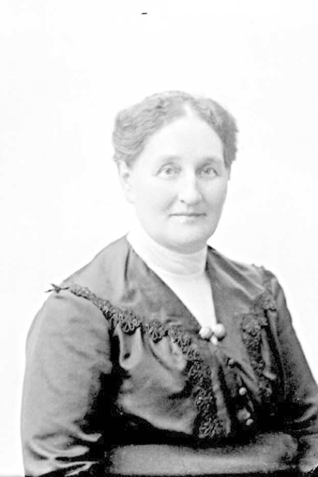 Schwarz-weißes Porträt einer selbstbewussten Frau in einem schlichten, eleganten Kleid mit langen Ärmeln und hohem Kragen, deren Haar zurückgebunden ist und die direkt in die Kamera blickt, mit einem warmen, bestimmten Ausdruck.