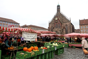 Ein belebter Markt in Nürnberg, Deutschland, mit verschiedenen Obst- und Gemüsesorten, Menschen, die umhereilen, Zelten, Gebäuden mit Fenstern und einem Uhrenturm im Hintergrund unter einem sichtbaren Himmel.