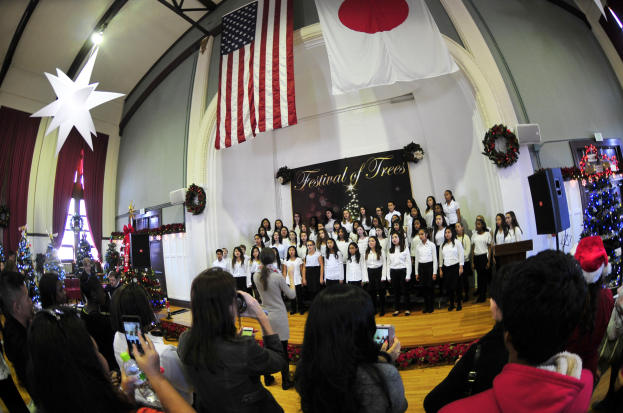 Gruppe von Menschen auf einer Bühne mit Weihnachtsdekorationen, einem Rednerpult, Lautsprechern und einer Tafel mit Text sowie einer Menge mit Kameras und Handys.