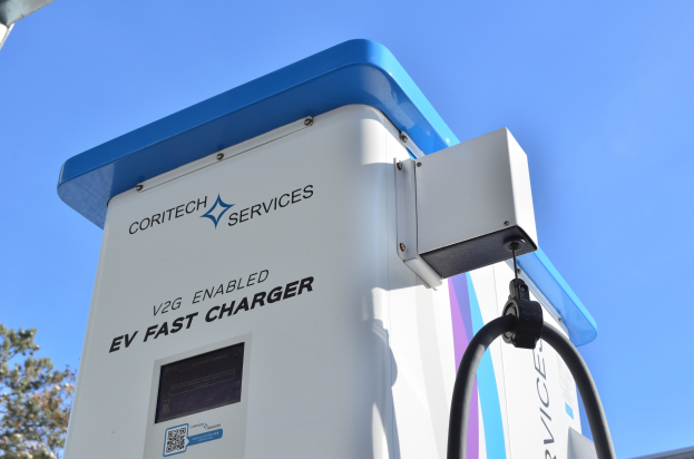 Elektrofahrzeug-Ladestation mit einem "V2G Fast Charger"-Schild, B├Ąumen, einem Geb├Ąude und einem klaren blauen Himmel.