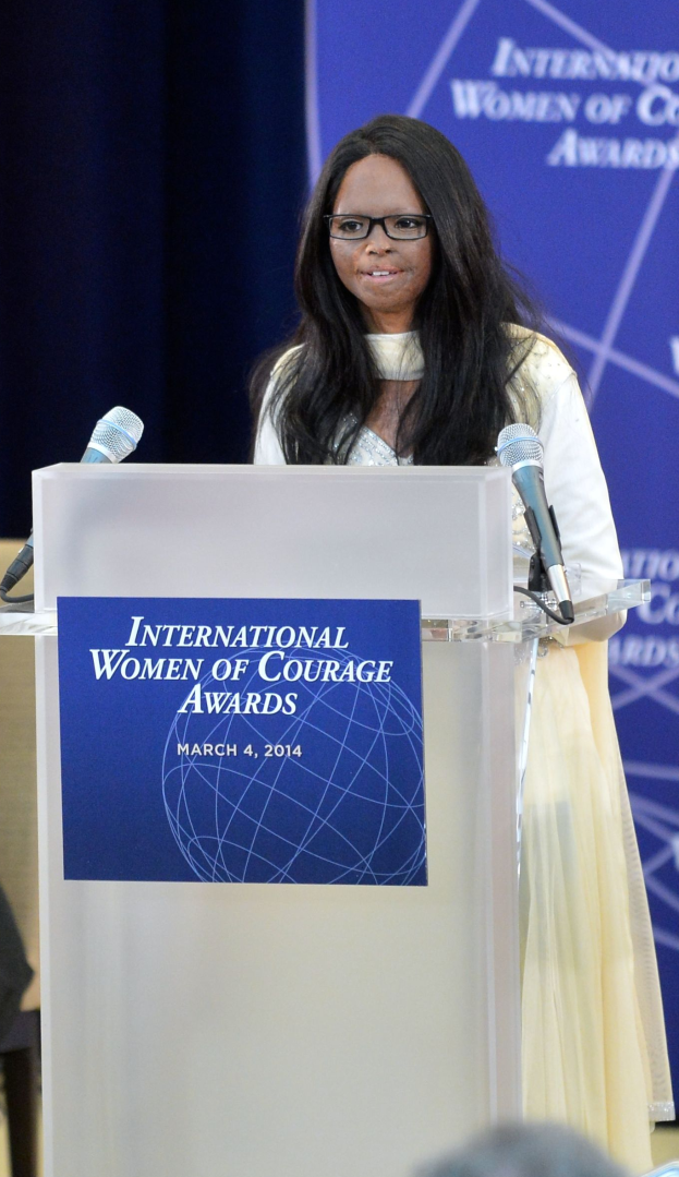 Eine Frau in einem weißen Kleid und Brille steht an einem Podium mit zwei Mikrofonen und einer Tafel, während sie eine Menge bei der Veranstaltung "International Women of Courage Awards 2014" anspricht, mit einem blauen Banner im Hintergrund.
