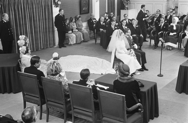 Schwarz-weißes Foto einer Hochzeitszeremonie mit sitzenden und stehenden Menschen, einem Mann mit Mikrofon, einer Frau mit Blumenstrauß, einem Podium mit Mikrofon, Blumenschmuck, einem Vorhang, einem gerahmten Foto an der Wand und einer Tür im Hintergrund.