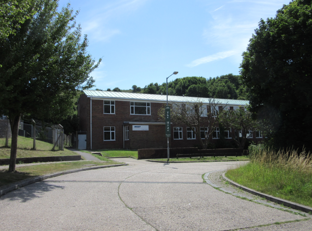 Ein Schulgebäude mit Fenstern, einem Straßenschild, einem Schild, einer Baumgruppe, Gras, einem Zaun und einem bewölkten Himmel, das in der Mitte eines Parks umgeben von saftigem Grün liegt.
