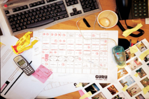 Ein unordentlicher Schreibtisch mit Papieren, einem Computer, einem Telefon, Tassen und einem Mobiltelefon.