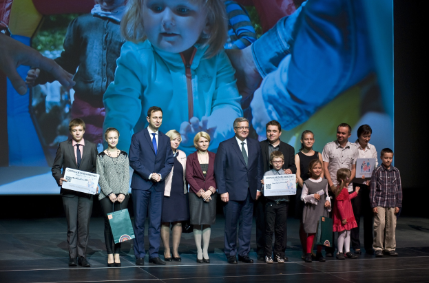 Eine Gruppe von Menschen auf einer Bühne mit Taschen und Urkunden, mit einem Bildschirm im Hintergrund, der ein Kind und andere zeigt, was auf ein Wohltätigkeitsereignis hinweist.