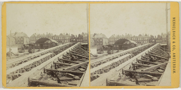 Schwarzes und weißes Foto einer Bahnschiene mit Gebäuden im Hintergrund, Text 'Merkelbach & Co, Co. Amsterdam' rechts und verstreute hölzerne Gegenstände im Vordergrund.