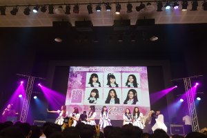 Eine Gruppe von Menschen performs auf einer hell beleuchteten Bühne mit Trägern, während die Menge darunter das Ereignis aufzeichnet; ein großer Bildschirm im Hintergrund zeigt Bilder der K-Pop-Gruppe BK48.