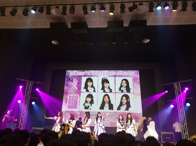 Eine Gruppe von Menschen performs auf einer hell beleuchteten Bühne mit Trägern, während die Menge darunter das Ereignis aufzeichnet; ein großer Bildschirm im Hintergrund zeigt Bilder der K-Pop-Gruppe BK48.