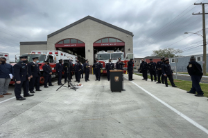 Gruppe von Menschen in Mützen neben einem Feuerwehrauto auf einem Podium während einer Feuerwachen-Einweihungszeremonie, mit Gebäuden, Bäumen, Strommasten und bewölktem Himmel im Hintergrund.