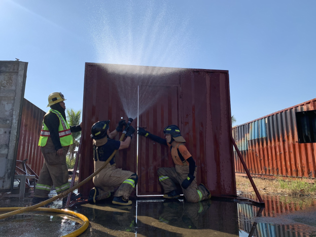 Feuerwehrleute in Helmen und Handschuhen spritzen Wasser aus einem Schlauch auf einen Container, um ein Feuer zu löschen, mit Bäumen und einem klaren blauen Himmel im Hintergrund.