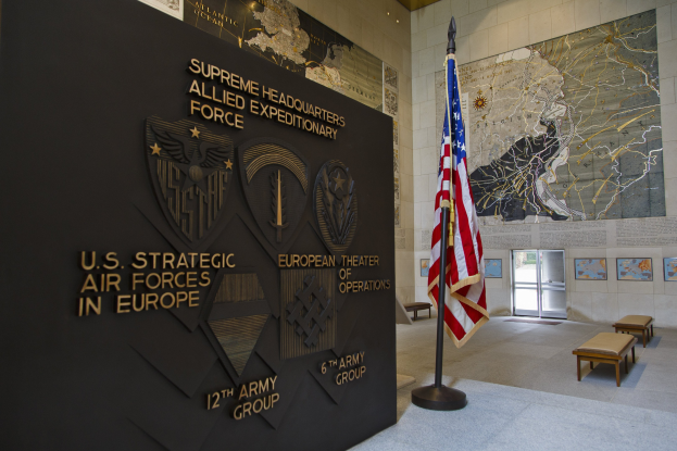 Eingang zum US-Strategischen Luftwaffenhauptquartier in Europa, mit einer amerikanischen Flagge, einer Wand mit Text und Logos, Bänken, einer Tafel, Fotorahmen, beleuchteter Decke und einer Tür im Hintergrund.