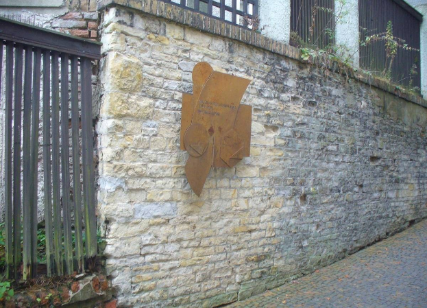 Steinmauer mit Metalltor auf der linken Seite, Gebäude mit Fenstern und Pflanzen im Hintergrund sowie eine Tafel an der Wand, die die Eröffnung des alten Rathauses erinnert.