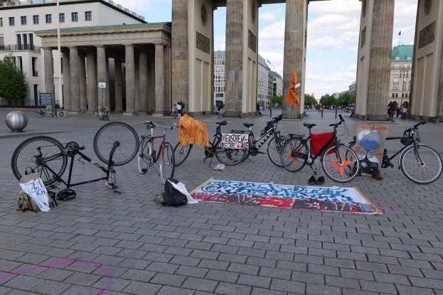 Eine Gruppe von Fahrrädern vor dem Brandenburgertor in Berlin, Deutschland, mit Taschen und einer Fahne in der Nähe, vor Gebäuden, Bäumen und einem klaren blauen Himmel.