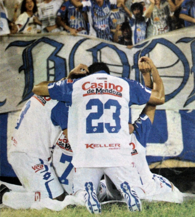 Eine Gruppe von Fußballspielern, die ein Tor auf dem Feld feiern, mit einer jubelnden Menge im Hintergrund und einem Banner mit Text dahinter.