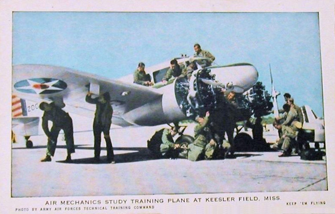 Eine Gruppe von Menschen um ein Trainingsflugzeug auf einer Landebahn mit einem klaren blauen Himmel im Hintergrund, beschriftet mit "Air Mechanics Study Training Plane at Keesler Field, Miss."