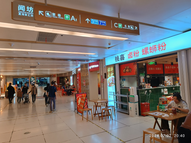 Eine Gruppe von Menschen geht durch ein Einkaufszentrum mit Tischen, auf denen verschiedene Gegenstände ausgelegt sind, Texttafeln an den Wänden, Deckenlampen und einem zentralen kreativen Markt mit Essensauslagen.