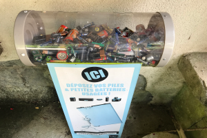 Ein Mülltonnen voller Batterien auf einem Bürgersteig, mit einem Plakat mit Text an der Wand dahinter und Stufen auf der rechten Seite.