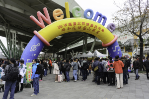 Eine Gruppe von Menschen vor einem Willkommensbogen bei einem Outdoor-Event, umgeben von Zelten, Bäumen und Gebäuden unter einem klaren blauen Himmel, wobei einige Teilnehmer Taschen tragen.