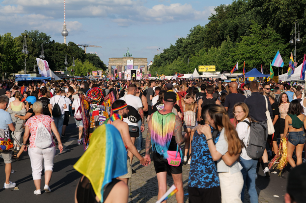 Eine große Menschenmenge geht eine Straße in Berlin, Deutschland, entlang, viele tragen Taschen und halten Fahnen, mit Zelten an den Seiten und Gebäuden im Hintergrund.