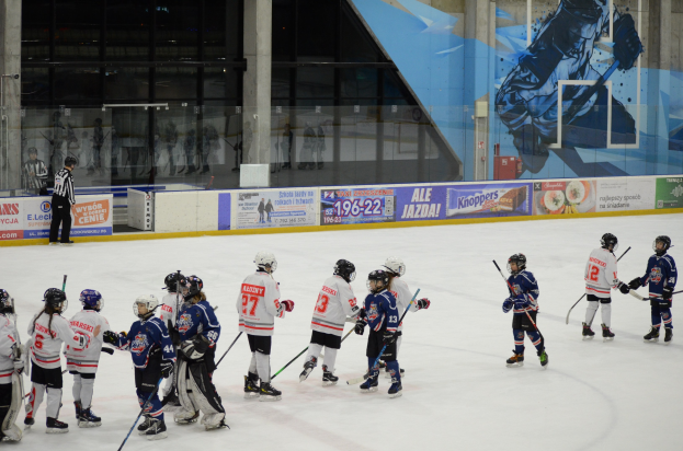 Eine Gruppe junger Menschen, die Eisockey auf einer Indoor-Eisbahn spielen, tragen Helme und halten Stöcke, mit einer Person links daneben und Bannern, Säulen und Glaswänden im Hintergrund.