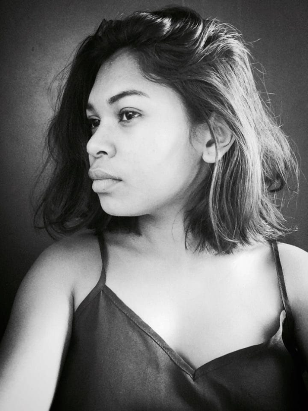 Eine selbstbewusste Frau in einem schwarzen Kleid posiert in einem Schwarz-Weiß-Foto, das an einer Wand mit einem glatten Bob und natürlichem Make-up steht.