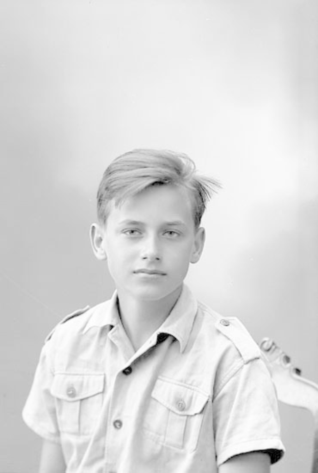 Ein junger Junge in einem Schwarz-Weiß-Foto hält eine Gitarre, trägt ein Hemd mit ernstem Gesichtsausdruck, kurze dunkle Haare und konzentrierte Augen auf dem Instrument.