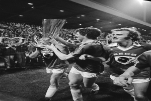 Schwarzes Bild von Fußballspielern, die mit einem Pokal auf dem Feld feiern, umgeben von einer Menge hinter einem Zaun unter Stadionbeleuchtung.