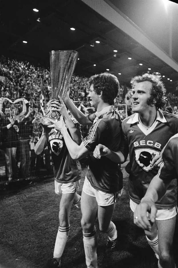 Schwarzes Bild von Fußballspielern, die mit einem Pokal auf dem Feld feiern, umgeben von einer Menge hinter einem Zaun unter Stadionbeleuchtung.