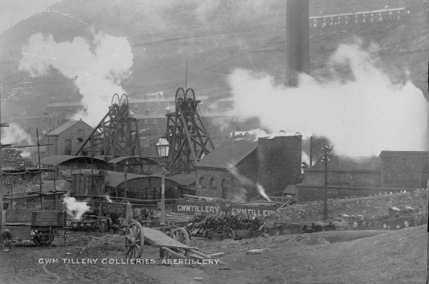 Schwarzes und weißes Foto einer rauchenden Fabrik mit umgebenen Gebäuden, Pfählen, Fahrzeugen, Karren und anderen Gegenständen; Text unten.